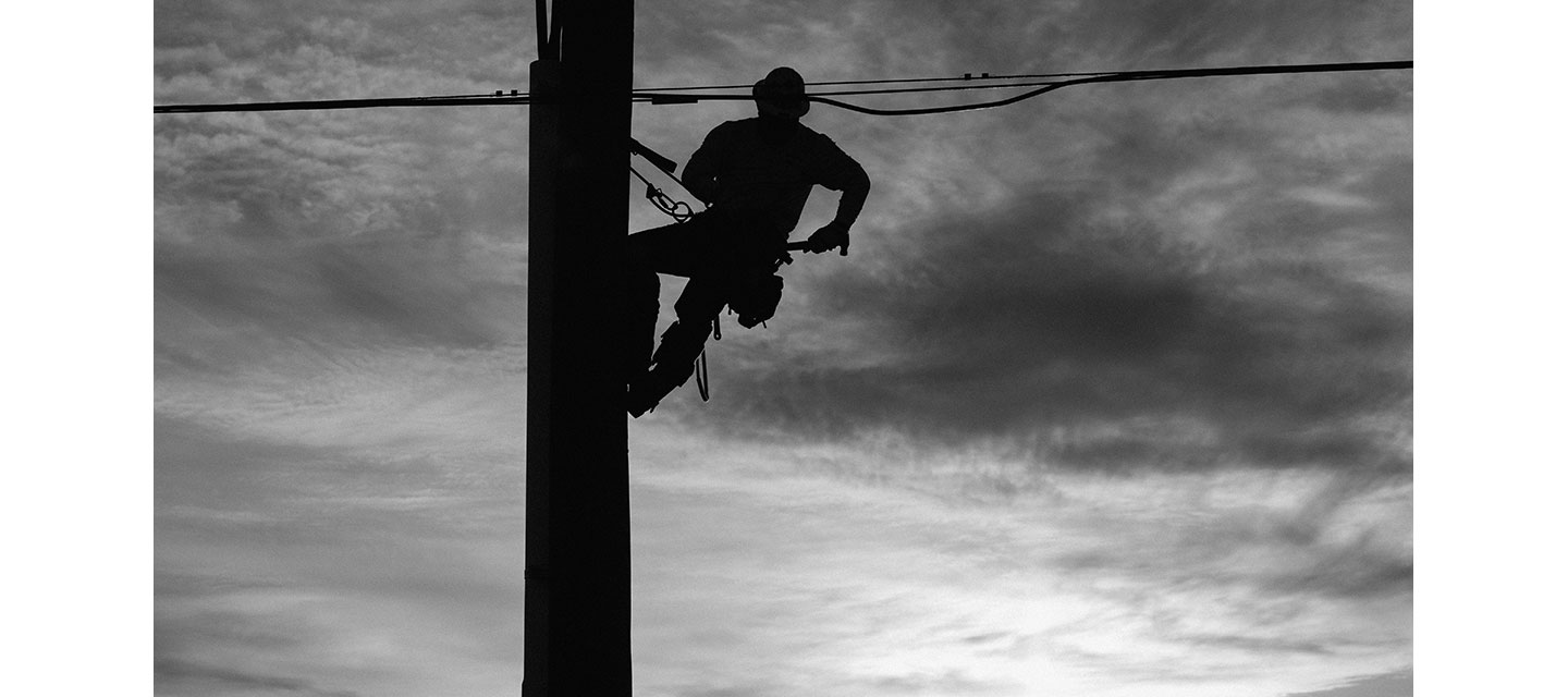 Una silueta de un liniero trepando un poste de servicios públicos con cables sobre su cabeza contra un cielo nublado.